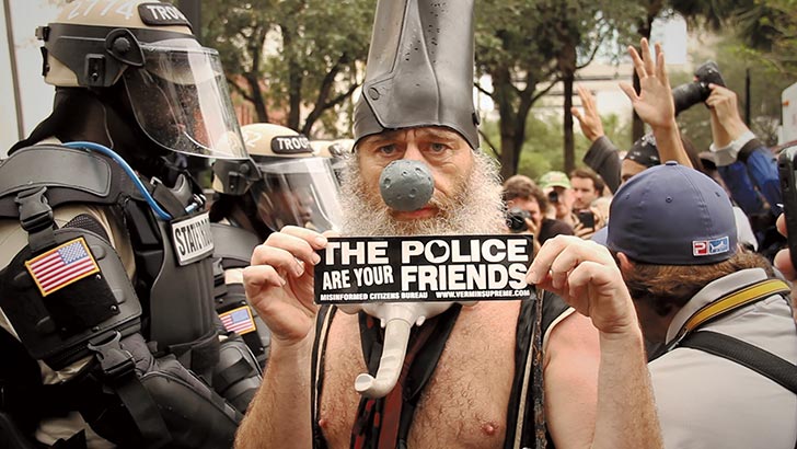 Vermin Supreme, shirtless holds up his "The police are your friends" in front of some heavily armored and armed friends.